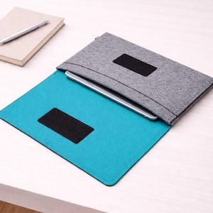 Puede incluir: Una funda gris de fieltro para tableta con un acento negro, abierta para revelar un interior turquesa y una tableta plateada. Un cuaderno y un bolígrafo están en el fondo. La funda está diseñada para proteger y transportar dispositivos electrónicos.