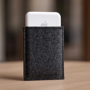 Könnte beinhalten: Eine dunkelgraue Filzhülle mit einem teilweise sichtbaren weißen elektronischen Gerät. Das Gerät hat eine abgerundete Oberseite und ein silbernes Apple-Logo. Die Hülle ist rechteckig und scheint aus einem weichen, strukturierten Material zu bestehen.