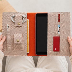 Peut inclure: Étui pour tablette à carreaux marron avec plusieurs poches et compartiments. L'étui est ouvert, révélant une tablette, un stylo, des câbles de chargement et un porte-cartes rouge. L'étui est tenu par une personne portant un pull crème.