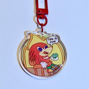 May include: A clear acrylic keychain featuring a cartoon character with red fur, holding a gem, with the text "THIS IS FINE" in a speech bubble. The keychain has a red clasp.