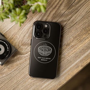 Peut inclure: Coque de téléphone noire avec un dessin blanc d'un bol de bortsch et le texte "KEEP CALM AND EAT BORSCHT". La coque est posée sur une surface en bois, avec un appareil photo et une plante en arrière-plan.