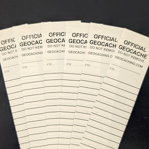 Puede incluir: Un abanico de tarjetas rectangulares blancas de « Official Geocache » con texto negro. Cada tarjeta tiene las palabras « OFFICIAL GEOCACHE » y « DO NOT REMOVE » impresas, junto con la dirección del sitio web. Las tarjetas también tienen líneas para escribir.