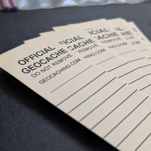 Puede incluir: Una pila de tarjetas rectangulares de color crema con texto negro. Las tarjetas dicen "OFFICIAL GEOCACHE" y "DO NOT REMOVE". Hay líneas para escribir debajo del texto. El sitio web geocaching.com también está impreso.