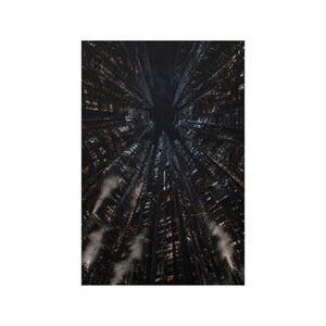 May include: An upward view of a city at night, with tall buildings reaching towards a dark sky. The buildings are illuminated with numerous lights, creating a grid-like pattern. Wisps of fog or clouds are visible near the base of the buildings.