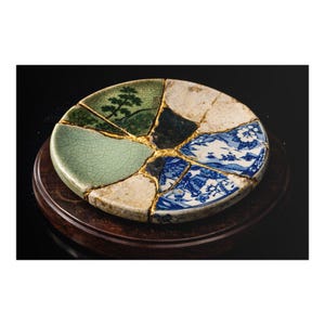 May include: A ceramic plate, meticulously reassembled using the Japanese art of Kintsugi. The plate's fragments, featuring green, white, and blue sections, are joined with gold. The plate is displayed on a dark wooden stand against a black backdrop.