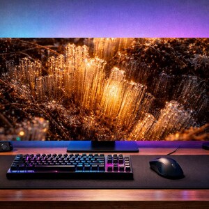 May include: A modern computer setup featuring a large monitor displaying an abstract, golden-toned image. A mechanical keyboard, mouse, and mousepad are in the foreground. A desktop computer, headphones, and plants complete the setup.