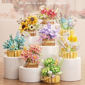 May include: A collection of eight small building block flower arrangements in various colors including yellow, pink, purple, blue, and green. Each set is displayed in a small basket-style container with a small tag reading Boutique.