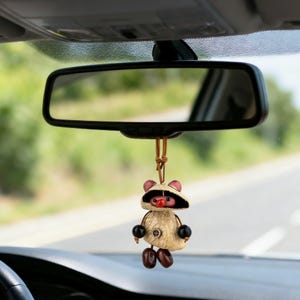 May include: A car rearview mirror with a decorative hanging ornament. The ornament is a tan-colored figure with a straw hat, black bead arms, and brown bean-shaped feet. The figure has a red nose and pink ears.