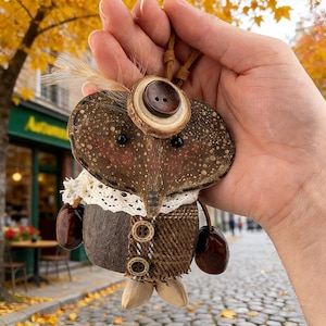 May include: Handmade whimsical owl pendant. The owl is crafted from natural materials, including wood, coconut shell, and fabric. It features a button eye, a feather, and a lace collar. The owl pendant is held by a brown leather cord.