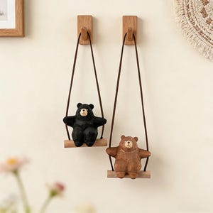 Puede incluir: Dos ganchos de madera montados en la pared, cada uno con un columpio colgante. Un columpio presenta una figura de oso negro y el otro una figura de oso marrón. Ambos osos tienen detalles tallados, hocicos claros y están sujetos por cordones.