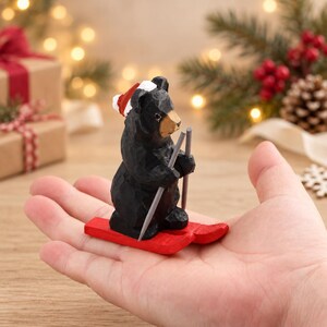 May include: A small, hand-carved black bear figurine wearing a red and white Santa hat. The bear is positioned on bright red skis and holds two grey ski poles. The figure features a textured, dark finish with a tan-colored snout.