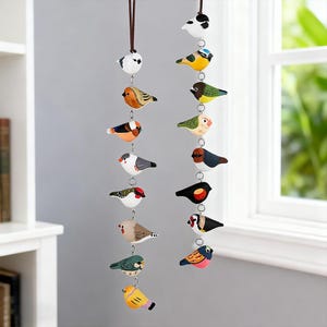 May include: Two strings of painted wooden bird decorations suspended from brown cords. The birds feature detailed patterns and colours, including blue, yellow, orange, and red. Small metal rings connect the birds, forming a decorative mobile for the home.