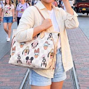 May include: A woman smiles while holding a large tote bag featuring a checkered pattern and cartoon character faces. The bag has a light-colored background with images of Mickey Mouse, Minnie Mouse, Donald Duck, and Goofy. She wears sunglasses, a white shirt, and denim shorts.