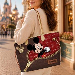 K&ouml;nnte beinhalten: Braune Tragetasche mit Mickey Mouse, die aus einem Rei&szlig;verschluss-Design hervortritt. Die Tasche hat ein rotes Innenfutter mit Mickey Mouse-Mustern und einen goldfarbenen Rei&szlig;verschluss. Ein Mickey Mouse-Anh&auml;nger h&auml;ngt an der Seite. Cremefarbene Griffe.