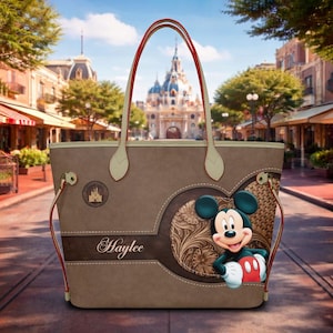 May include: A brown tote bag featuring a Mickey Mouse design. The bag has red handles and a decorative heart-shaped element with a floral pattern. The name "Haylee" is printed on the bag.