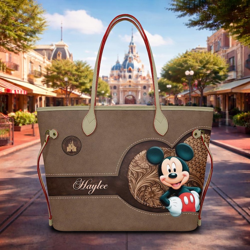 May include: A brown tote bag featuring a Mickey Mouse design. The bag has red handles and a decorative heart-shaped element with a floral pattern. The name "Haylee" is printed on the bag.