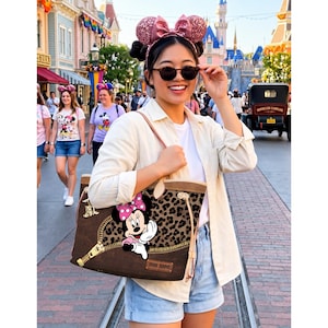 Op de afbeelding: Een bruine tote bag met een luipaardprint en een Minnie Mouse-afbeelding. De tas heeft een ritssluiting en een lichtbruine rand. De tas wordt gedragen door een persoon met een zonnebril en een wit overhemd.