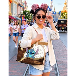 Puede incluir: Un bolso tote marr&oacute;n y beige con un personaje de dibujos animados Stitch asomando por un dise&ntilde;o de cremallera. El bolso tiene un fondo blanco con un patr&oacute;n de Stitch y una etiqueta de nombre personalizable. El bolso lo lleva una persona en un parque tem&aacute;tico.