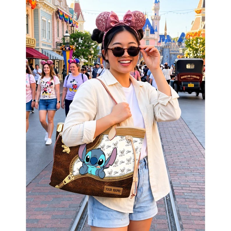 Puede incluir: Un bolso tote marr&oacute;n y beige con un personaje de dibujos animados Stitch asomando por un dise&ntilde;o de cremallera. El bolso tiene un fondo blanco con un patr&oacute;n de Stitch y una etiqueta de nombre personalizable. El bolso lo lleva una persona en un parque tem&aacute;tico.