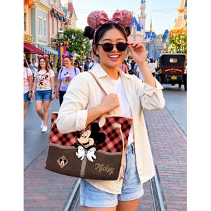 May include: A red and black plaid tote bag featuring Mickey Mouse. The bag has a brown base with a Mickey Mouse patch and the name "Mickey" printed on it. The bag is carried by a person wearing sunglasses and a white shirt.