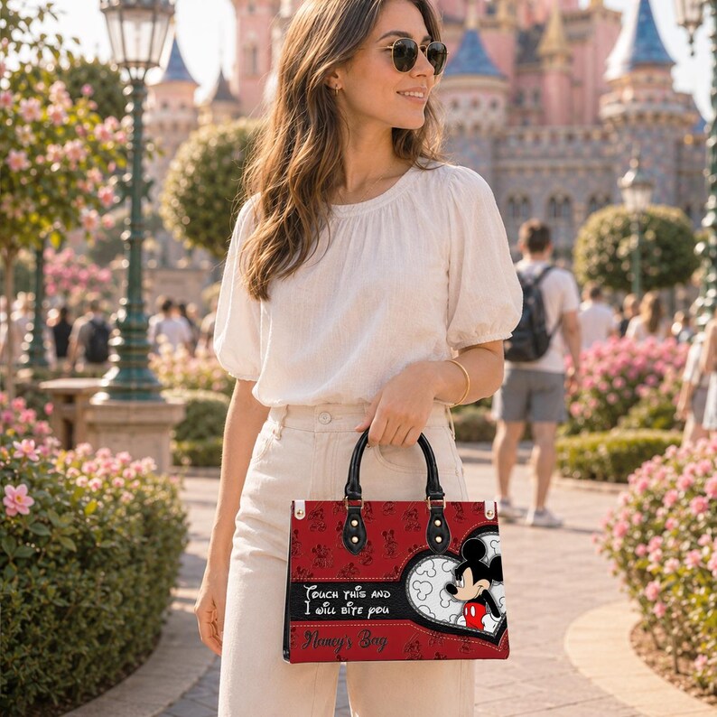 May include: A red rectangular handbag featuring a repeating Mickey Mouse pattern, a large heart-shaped graphic with Mickey Mouse, and the text "Touch this and I will bite you" followed by "Nancy's Bag" in black script on the front panel.