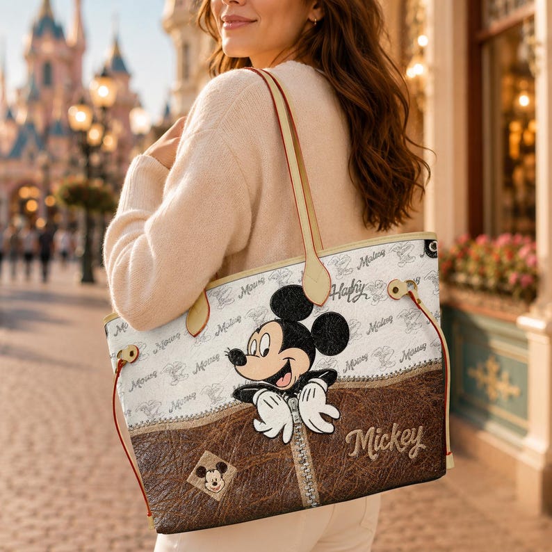 May include: A cream-colored tote bag featuring Mickey Mouse. The bag has a brown leather-like panel with a zipper design and the name "Mickey" embroidered. The bag has cream-colored handles and a repeating pattern of "Mickey" in a script font.