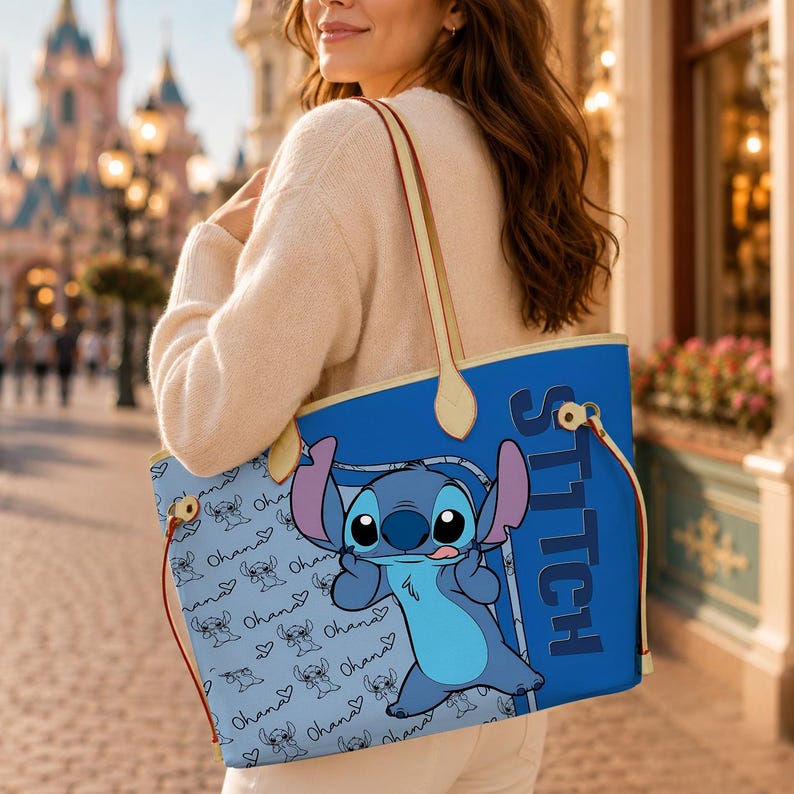 May include: A blue tote bag featuring the character Stitch from Lilo & Stitch. The bag has a light blue background with Stitch and "Ohana" patterns. The word "Stitch" is printed vertically on the side. Tan straps and trim complete the design.
