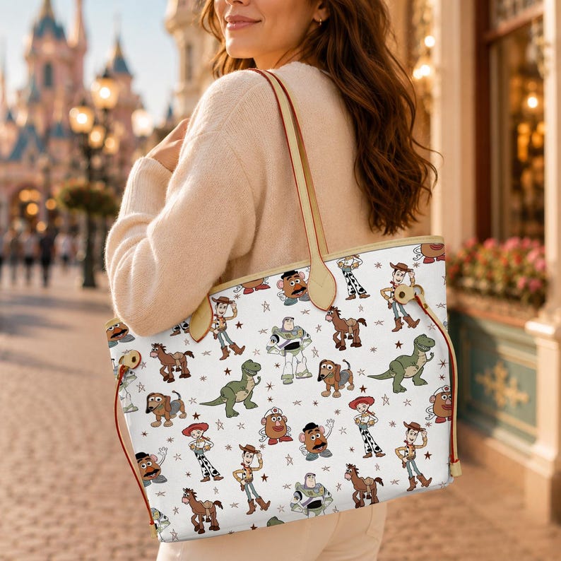 May include: White tote bag featuring a repeating pattern of Toy Story characters, including Woody, Buzz Lightyear, and Mr. Potato Head. The bag has tan straps and trim, with red accents. The bag is being carried by a person.