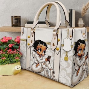 May include: White handbag featuring a Betty Boop cartoon design. The bag has a rectangular shape with dual handles, gold-colored hardware, and a chain detail. The Betty Boop graphic is repeated on both sides of the bag.