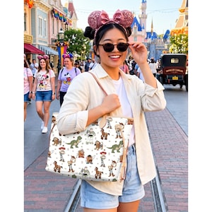 May include: A white tote bag featuring a pattern of Toy Story characters. The bag has brown leather handles and trim. The background shows a theme park with people and a castle.