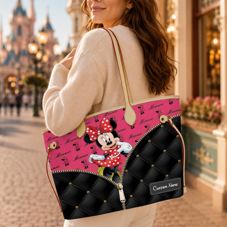 May include: A pink tote bag featuring Minnie Mouse in a red polka dot dress and bow. The bag has a black quilted leather-like design with a zipper detail and gold-colored hardware. The bag has beige handles and the words "Minnie" are printed on the pink background.