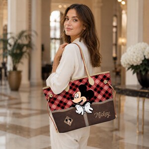 May include: A tote bag featuring a red and black plaid pattern with a brown base. The bag has a Mickey Mouse graphic and the word "Mickey" printed on the front. The bag has tan straps.
