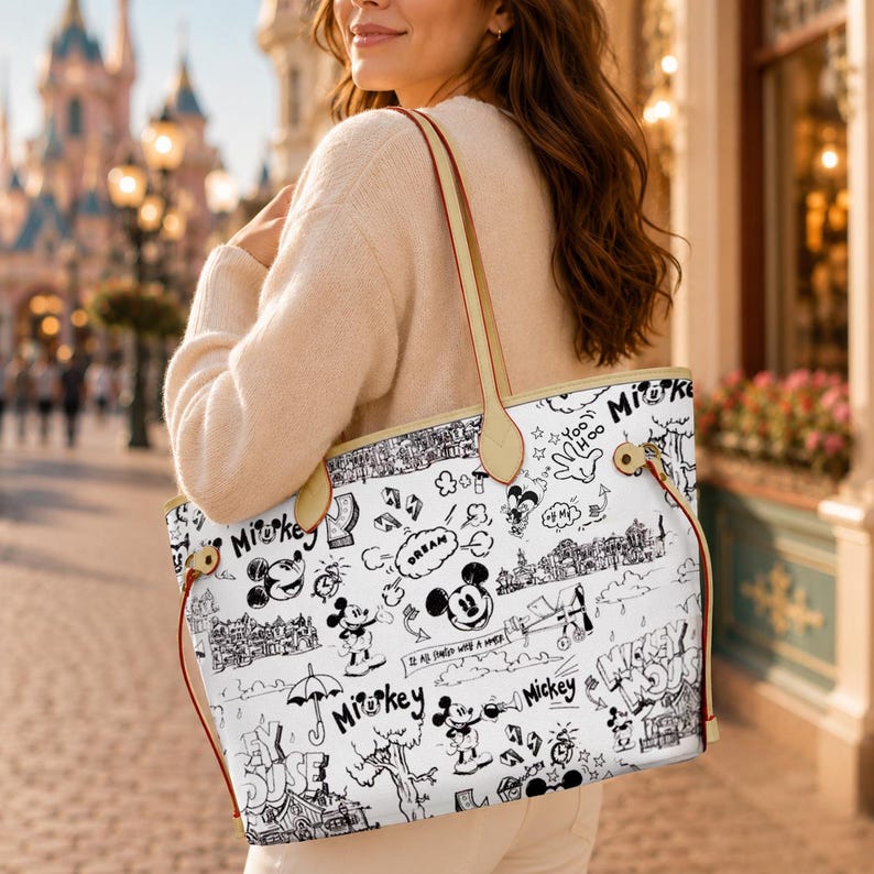 May include: White tote bag with a black and white Mickey Mouse doodle design. The bag has tan handles and red accents. The bag is carried over the shoulder.