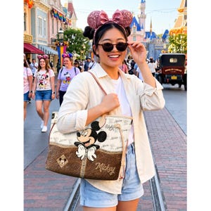 May include: A beige and brown tote bag featuring Mickey Mouse. The bag has a brown leather-like bottom section and a beige top section with a repeating pattern. The word "Mickey" is written in cursive. The bag is carried by a person wearing sunglasses.