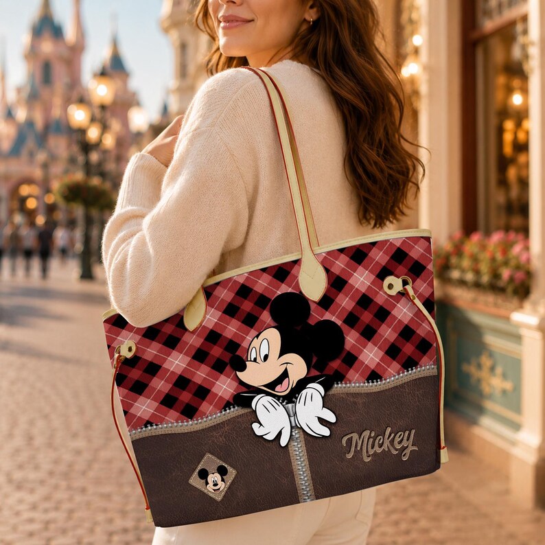 May include: A tote bag with a red and black plaid pattern, featuring a brown pocket with a zipper and the word "Mickey". The bag has a Mickey Mouse graphic and cream-colored handles.