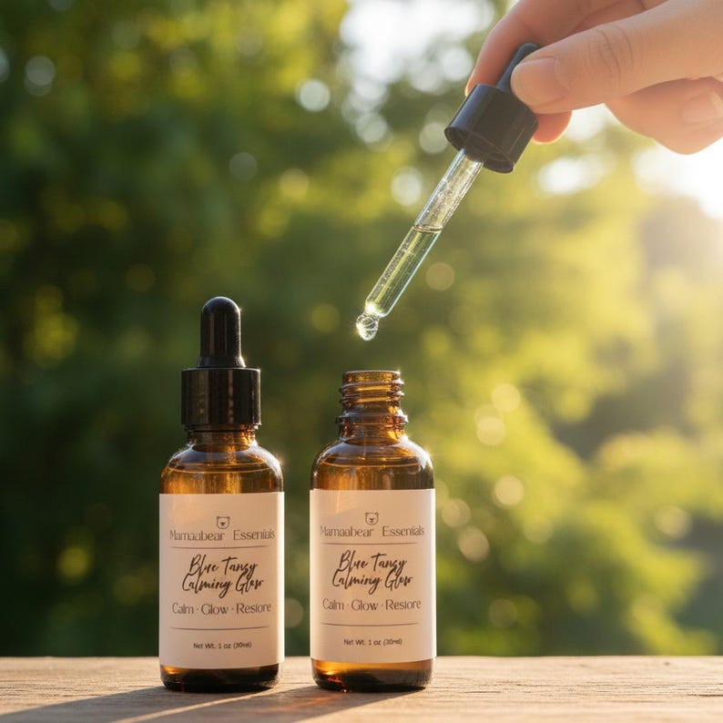 May include: Two amber glass bottles with droppers, labeled "Blue Tanzy Calming Glow." A dropper dispenses liquid into one bottle. The bottles are on a wooden surface with a blurred green background. Each bottle contains 1 fl oz.