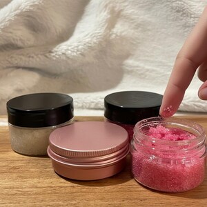 May include: An assortment of skincare products, including three small glass jars with black lids and a small pink tin. One jar contains pink scrub, and another contains a light-colored scrub. The products are on a wooden surface.