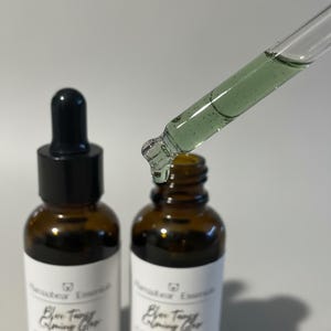 May include: Two amber glass bottles with black droppers, one with a dropper dispensing a green liquid. The bottles have white labels with black text. The product is likely a skincare serum or oil.