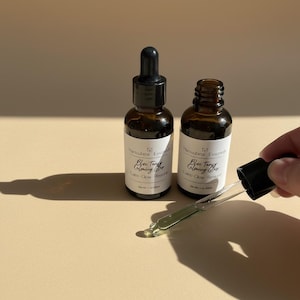 May include: Two brown glass bottles with black droppers and white labels. The labels read "Blue Tangy Calming Glow". A dropper is held up, filled with a clear liquid. The background is a neutral beige.