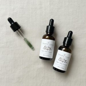 May include: Two brown glass bottles with black droppers and white labels that read "Blue Tangy Calming Glow." A glass dropper with green liquid is next to the bottles. The bottles are on a light beige surface.