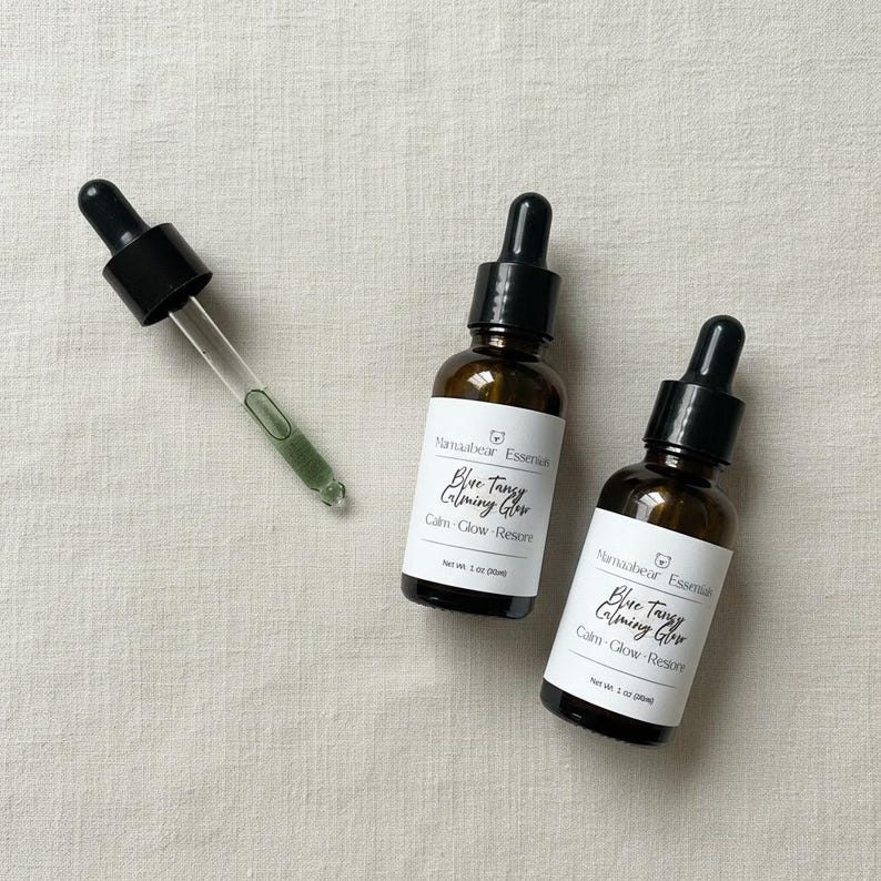 May include: Two brown glass bottles with black droppers and white labels that read "Blue Tangy Calming Glow." A glass dropper with green liquid is next to the bottles. The bottles are on a light beige surface.