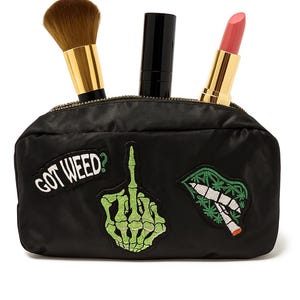 Puede incluir: Bolsa de cosméticos negra con cremallera dorada, con una brocha de maquillaje, lápiz labial y un tubo negro. La bolsa tiene parches con el texto "GOT WEED?" y una mano de esqueleto haciendo la peineta.