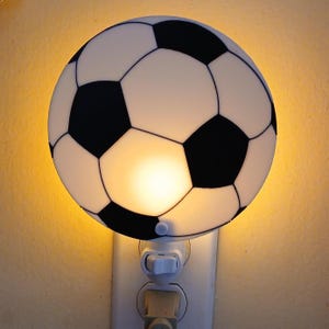 May include: A soccer ball-shaped night light with black and white panels. The light is plugged into a white electrical outlet and emits a warm glow. The night light is a decorative item for a child's room or sports-themed space.