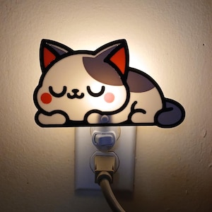 May include: A white and gray cat-shaped night light plugged into a wall outlet. The cat has closed eyes, a small mouth, orange inner ears, and pink circular cheeks. The light emits a soft glow from behind the cat's silhouette, highlighting its outline.