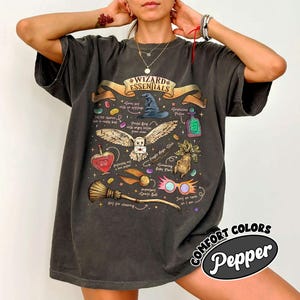 May include: A charcoal gray short-sleeve t-shirt featuring a graphic print titled Wizard Essentials. The design includes illustrations of a wizard hat, an owl with a letter, a potion bottle, a broom, a mandrake plant, and pink glasses with text.