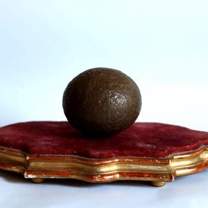 Puede incluir: Una esfera marrón oscuro y texturizada descansa sobre una plataforma de terciopelo rojo con un marco de madera dorado y ornamentado. La plataforma tiene un borde festoneado y está sobre un fondo blanco.