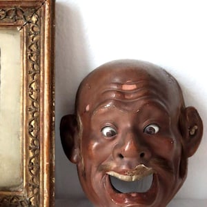 Könnte beinhalten: Eine Vintage-Maske in Brauntönen mit offenem Mund und ausdrucksstarken Augen. Die Maske hat einen kahlen Kopf, einen Schnurrbart und große Ohren. Links von der Maske befindet sich ein gerahmtes Bild mit einem goldfarbenen, verzierten Rahmen.