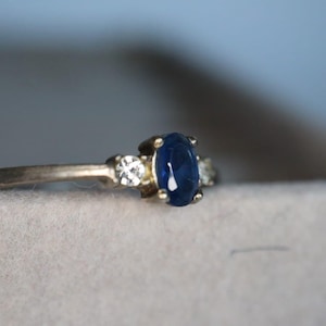Könnte beinhalten: Ein silberner Ring mit einem großen, ovalen, tiefblauen Edelstein als Mittelpunkt, flankiert von zwei kleineren, klaren Edelsteinen. Der Ring hat ein zartes Band und ein klassisches Design.