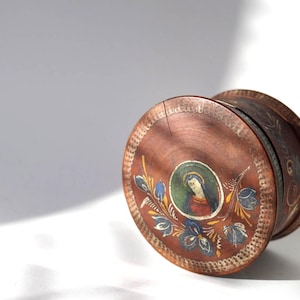 Könnte beinhalten: Eine runde Holzschatulle mit dunkelbraunem Finish, verziert mit einem handgemalten kreisförmigen Porträt einer Figur mit blauem Schleier, umgeben von Blumenmotiven. Der Deckel weist einen vertikalen Riss und dekorative weiße Malereien auf.