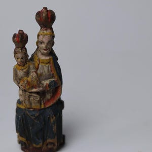 Pode incluir: Uma estatueta de madeira esculpida &agrave; m&atilde;o representando uma figura segurando uma crian&ccedil;a, ambas usando coroas. A escultura apresenta um acabamento de pintura desgastado em tons de azul, amarelo, vermelho e branco, com sinais de desgaste.